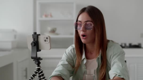 若い女性ブロガーは三脚の上にスマートフォンを見て、現代的なインテリアの背景にすごいと言う — ストック動画