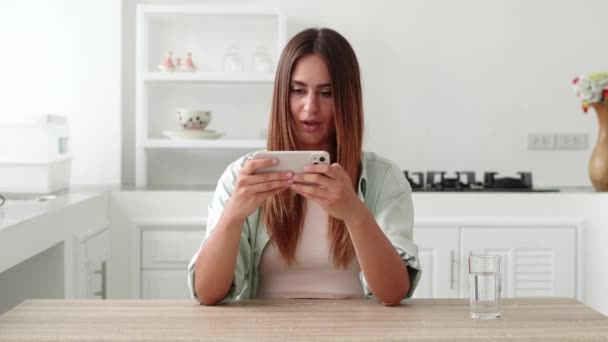 Jovem de cabelos castanhos mulher se divertindo jogando telefone. Imagens de stock. Jovem elegante divertido e entusiasticamente jogar jogos em seu smartphone sentado na cozinha moderna. Conceito de vitória — Vídeo de Stock