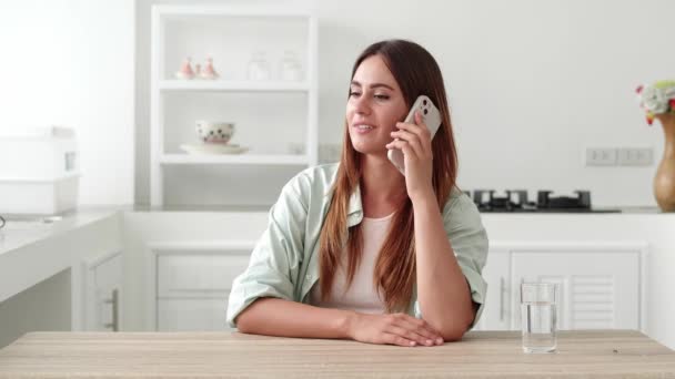 Vonzó 20-29 éves üzleti hölgy beszélni okos telefonon ügyfél, miközben ül az asztalnál a hazai konyhában. — Stock videók