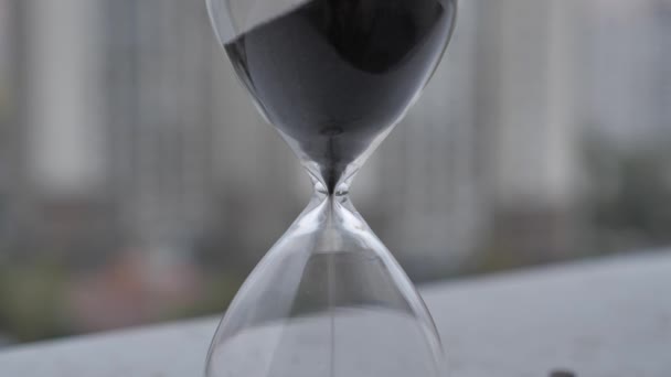 ブラーダイヤルの背景に黒い砂を流れる透明な砂時計の極端なクローズアップ 古典的なタイマー 時間概念 — ストック動画