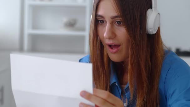 Привлекательная женщина в голубой рубашке сидеть на кухне держать в руках простыни читать отличные новости в письме чувствует себя счастливым праздновать успех. Фокус на бумаге — стоковое видео