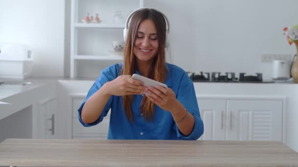 Красива дівчина сидить на кухні. Жінка в навушниках розслабляється, граючи в гру на своєму смартфоні, посміхаючись — стокове відео