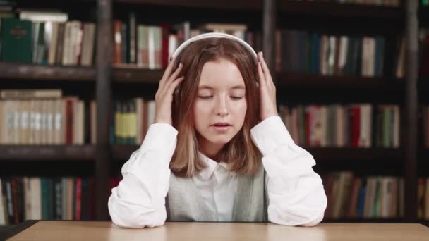 A beautiful small teen female in a library while listening to music with headphones and daydreams dancing to the music. Concept: educational, portrait, library, dancer — Vídeo de Stock