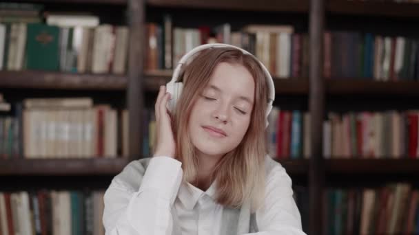 Adolescente com cabelo curto vestindo suéter rosa ouvindo música favorita com fones de ouvido enquanto sentado na biblioteca — Vídeo de Stock