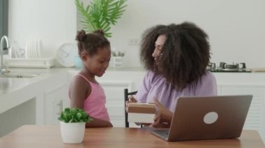 Charming African multitasking woman gently hugging her beloved daughter while opening the gift box for Mothers Day