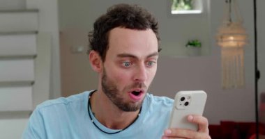 Handsome guy has fun at home, expressing surprised emotion while scrolling news feed and checking social media content