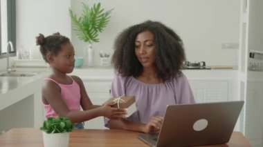 Multitasking woman works remotely on laptop, and rejoices while her daughter presenting her a gift for Mothers Day