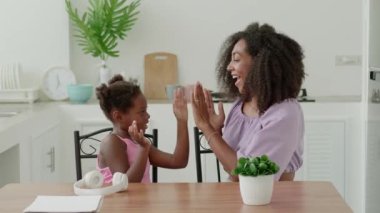 African pretty woman plays games with her cute daughter, claps hands, having fun together at cozy light home interior.