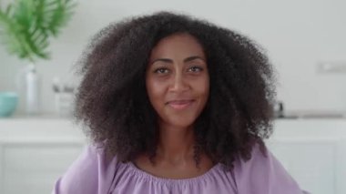 African pretty woman smiling a cheerful toothy smile while looking at camera, standing against a home kitchen background