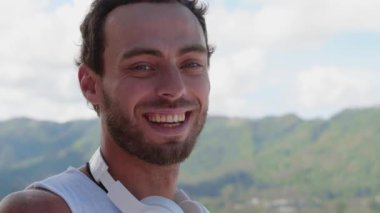 Headshot. Delighted young man tourist, with hands free audio headset, smiles at camera while admiring beautiful nature