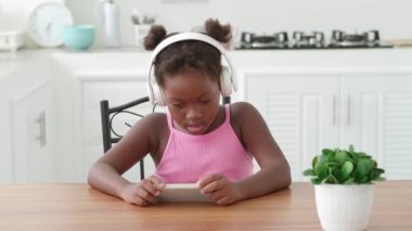 Cute little girl, in audio headset, using mobile phone, listens to the music, watching cartoons while relaxing at home