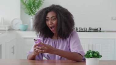 Charming positive woman, relaxing at home, using mobile phone, checking content and rejoices, receiving good news