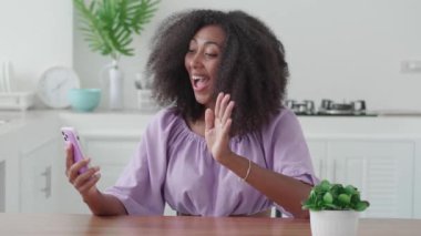 Pleasant young African woman in purple wear, having a video call while relaxing in the cozy home interior