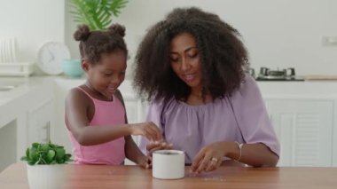 Delightful young loving mother helps her little daughter in saving money by collecting coins into piggy bank together.