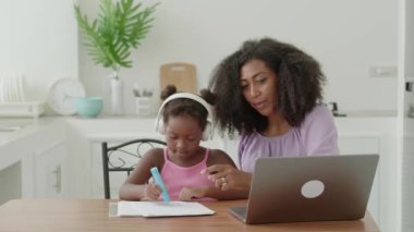Adorable kid girl in wireless headphones browsing drawing online lesson on laptop, sitting next to her loving mother