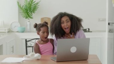 Astonished mom and daughter watching movie on laptop, expressing surprise and smiling together looking at each other.