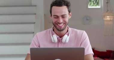 Caucasian man, sitting at wooden desk and works remotely on laptop, cheerfully laughing looking at the screen monitor