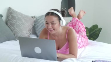 Güzel bir ev içinde beyaz bir yetişkin kadın. Dizüstü bilgisayarda yazan ve yatakta yatarken müzik dinleyen bir kız.