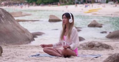 Asyalı kız nefes egzersizleri yapıyor ve plaja yakın meditasyon için rahatlatıcı müzik dinliyor.
