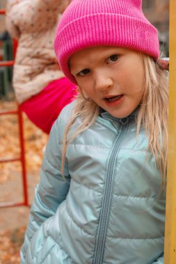 Hoş, komik, sarışın anaokulu kızı sonbahar günü dışarıda surat yapıyor. Pembe bereli ve mavi ceketli bir kız. Dikey çekim