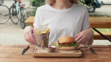 a girl in a white t-shirt takes a fork and a knife in her hands and cutting half of a hamburger on rusty wooden table. fast food concept