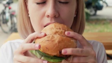 Close-up of a hungry girl eating a big hamburger outdoors. Joyful woman enjoying fast food dinner