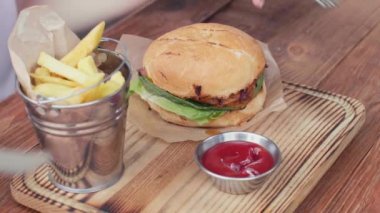 a girl takes a fork and a knife in her hands and cutting half of a hamburger on rusty wooden table. fast food concept. close-up