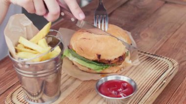 a girl takes a fork and a knife in her hands and cutting half of a hamburger on rusty wooden table. fast food concept. close-up