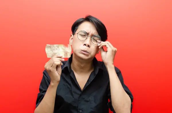 Asian young man with sad expression holding small amount of rupiah.