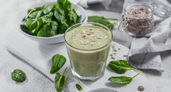 Стакан домашнего здорового зеленого смузи со свежим детским шпинатом, подсолнухами и семенами кунжута на светлом фоне. Еда и напитки, диета и здоровое питание