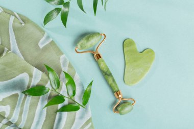 Top view of Green jade face roller and gua sha scraper with leaves on light blue background. Massage tool for facial skin care, SPA self and beauty care treatment concept.