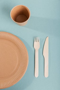 Tek kullanımlık kağıt sofra takımı. Bardaklar, tabaklar, ahşap çatallar ve mavi arka planda bıçaklar. Plastiksiz ve sıfır atık kavramı.