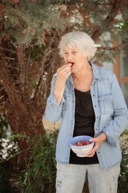 Elinde bir kase olgun çilek tutan ve tatlı meyveler yiyen yaşlı bir kadın.