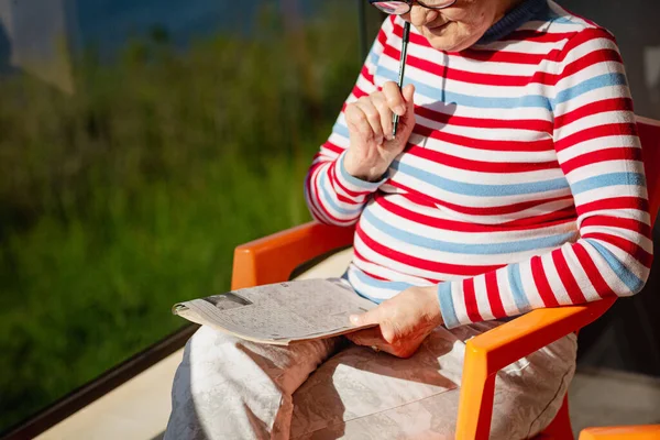 Elderly unrecognizable woman in glasses sitting on a balcony near the sea and looking at magazine, solving a crossword