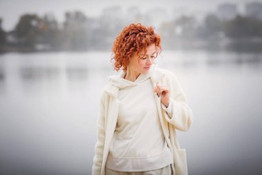 Beyaz, sıcak palto giymiş, elinde sarı yapraklar olan çekici bir kadın nehrin kenarında yürüyor. Sonbahar moda konsepti