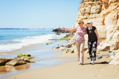 İki yaşlı kadın kayalık sahilde yürüyor, konuşuyor ve gülüyor, birlikte hoş vakit geçiriyorlar.