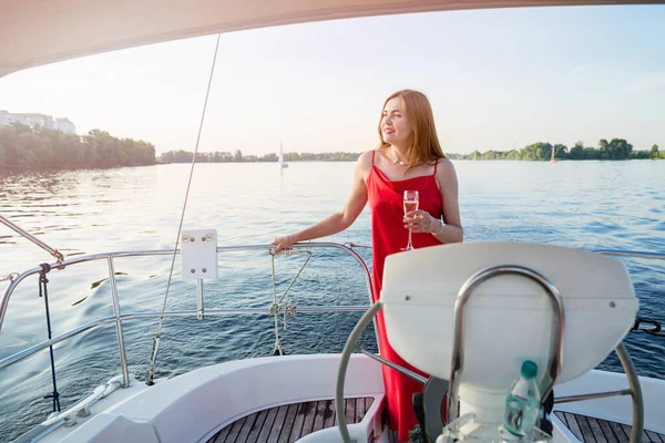 Elegant woman in a long red dress holding a glass of champagne standing on a yacht and looking for river sunset view