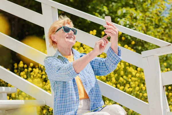 Mature attractive woman traveler sitting alone on the terrace of beach coffee shop in Bulgaria and using mobile phone. Active life of the elderly in retirement, active seniors