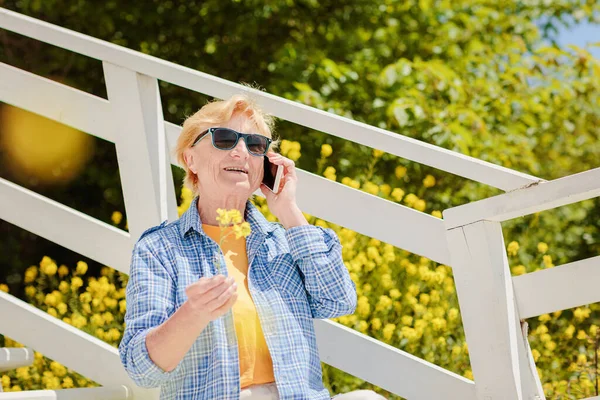 Mature attractive woman traveler sitting alone on the terrace of beach coffee shop in Bulgaria and using mobile phone. Active life of the elderly in retirement, active seniors