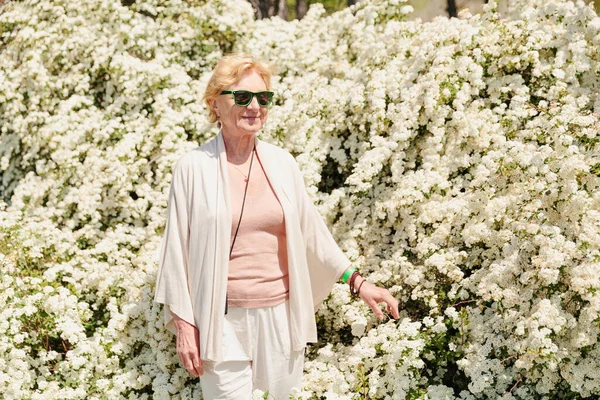 Starsza Kobieta Pozująca Wśród Krzewów Białymi Kwiatami Aktywni Seniorzy — Zdjęcie stockowe