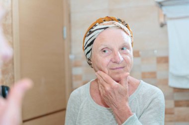 Olgun bir kadın yüzüne bakıyor. Banyoda aynanın karşısında duran ve yüzüne krem ya da maske süren son sınıf öğrencisi kadın.