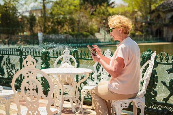 Mature attractive woman traveler sitting alone on the terrace of coffee shop in beautiful park in Bulgaria and using mobile phone. Active life of the elderly in retirement, active seniors