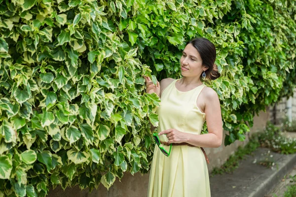 Woman staying surrounded by lush green plants, touching leaves. Caucasian woman near leaves wall, wearing yellow dress. Nature concept