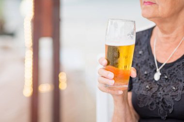 Unrecognisable Senior woman sitting at a table in a summer cafe and drinking beer from a tall glass