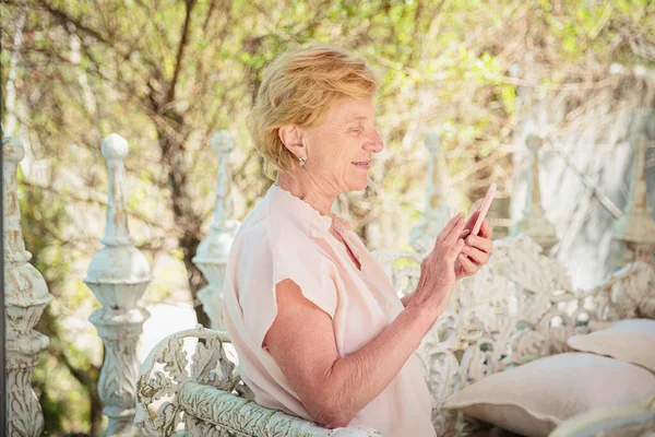 Mature attractive woman traveler sitting alone on the terrace of coffee shop in beautiful park in Bulgaria and using mobile phone. Active life of the elderly in retirement, active seniors