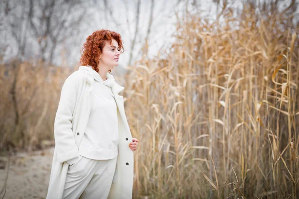 Güzel, beyaz elbiseli genç bir kadın, sonbahar manzarasının arka planında kuru sazlıklarla hareket ediyor.