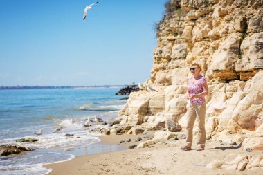 Güneş gözlüklü yaşlı kadın sahilde martıları besliyor ve gülüyor. Mutlu emeklilik hayatı