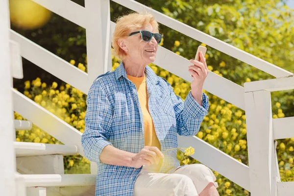 Mature attractive woman traveler sitting alone on the terrace of beach coffee shop in Bulgaria and using mobile phone. Active life of the elderly in retirement, active seniors