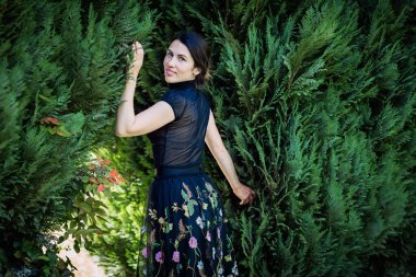 A young pretty woman in black clothes is standing near a bush. Photography in a dark key