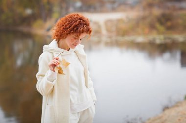 Beyaz, sıcak palto giymiş, elinde sarı yapraklar olan çekici bir kadın nehrin kenarında yürüyor. Sonbahar moda konsepti